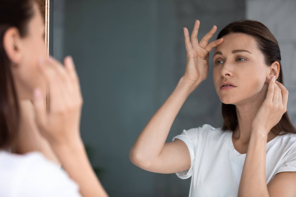 Woman examining forehead lines in mirror, considering brow lift to fully remove wrinkles.